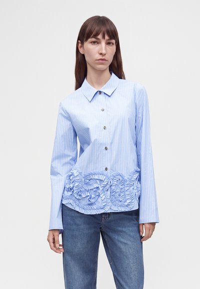 Femme portant une chemise rayée bleu clair à manches longues avec une broderie florale à volants en bas, associée à un jean bleu, sur fond blanc.