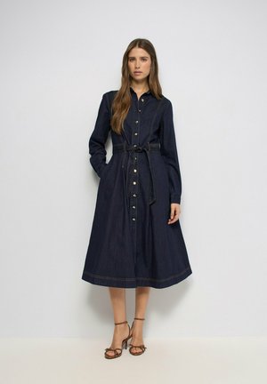 Mujer vestida con un vestido camisa azul oscuro abotonado, con mangas largas y cinturón atado en la cintura, lleva sandalias marrones de tacón alto con tiras.
