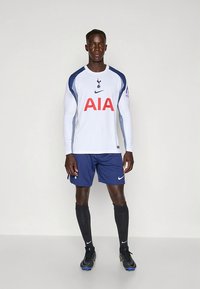 Maillot de football blanc à manches longues avec accents bleus, arborant un grand logo rouge "AIA" et un emblème du club, associé à un short bleu marine et des chaussettes noires.