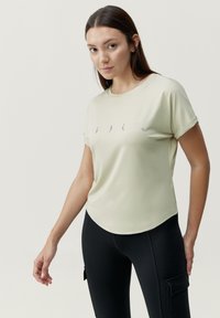Lichtgroene top met korte mouwen en een ronde halslijn, met een gebogen zoom, voorzien van een eenvoudige grijze grafische afbeelding op de borst, gedragen met zwarte leggings.