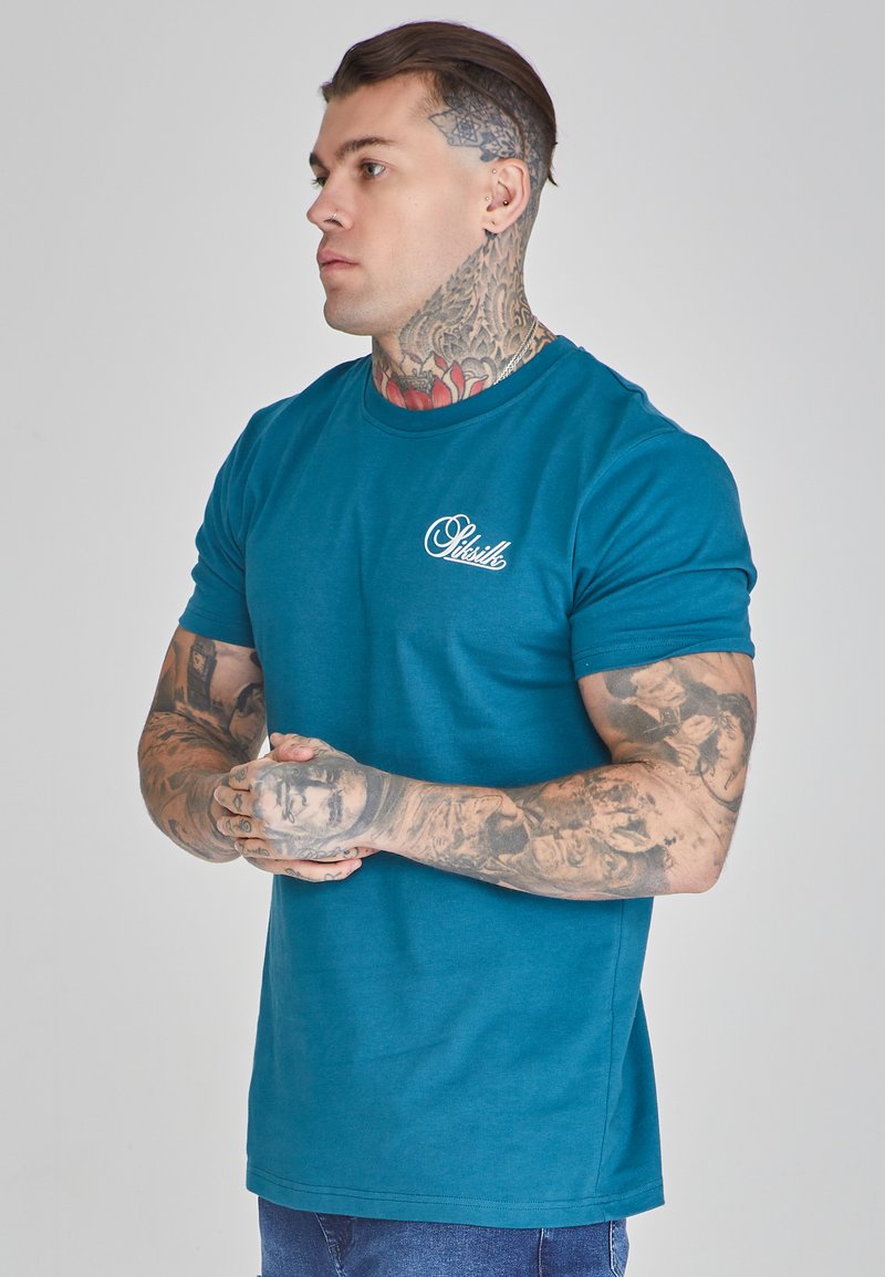 Türkises Baumwoll-T-Shirt mit kurzen Ärmeln, runder Ausschnitt und einem weißen Logo auf der linken Brust; das Modell hat sichtbare Tattoos und ist in einer neutralen Pose.