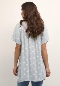 Tunique bleu clair à manches courtes, en tissu texturé, avec un motif floral blanc. Sa longueur atteint le mi-thigh et elle a une coupe décontractée.