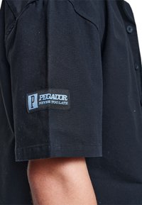 Camisa negra de manga corta hecha de algodón, con cierre de botón y un parche de logo en la manga izquierda con "Pegador Never Too Late."