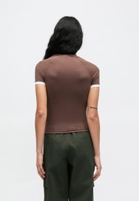 Καφέ κοντομάνικη μπλούζα με εφαρμοστή γραμμή, διακοσμημένη με λευκή ρίγα στο λαιμό και τα μανίκια. Φορεμένη με πράσινο παντελόνι cargo.