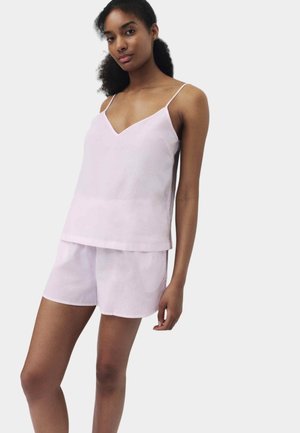 Femme portant un haut sans manches rose clair à col en V et un short assorti, debout sur un fond uni.