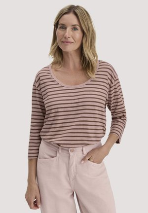 Frau mit schulterlangem blondem Haar trägt ein gestreiftes Hemd in Mauve und Braun sowie helle rosa Hosen, steht mit einer Hand in der Tasche.