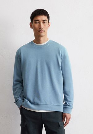 Light blue crewneck sweatshirt made of soft fabric with ribbed cuffs and hem, layered over a white shirt, set against a plain background.