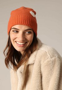 Oranje gebreide beanie met een ribbelstructuur en een witte hartaccent. Draagt men met een crèmekleurige nep-bontjas, met een comfortabele uitstraling.