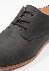 Chaussure en suède noir avec un bout rond, un laçage à l'avant, des coutures minimales et une semelle brun clair. Texture lisse avec une finition mate.