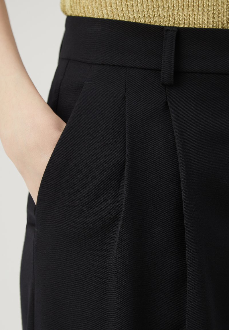 Zwarte op maat gemaakte broek van textuurstof, met geplooide voorkant en zijzak, gecombineerd met een geribbeld gouden top.