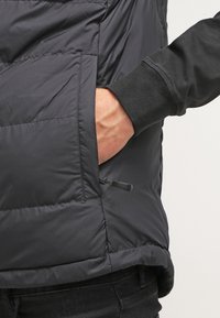 Svart pufferväst med sidoficka, försedd med dragkedja och en slät yta. Inre lagret är en kontrasterande svart långärmad skjorta.