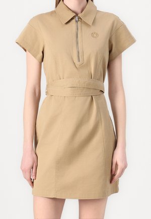 Robe chemise beige à manches courtes avec col, fermeture éclair devant et ceinture à la taille portée par une personne debout devant un fond uni.