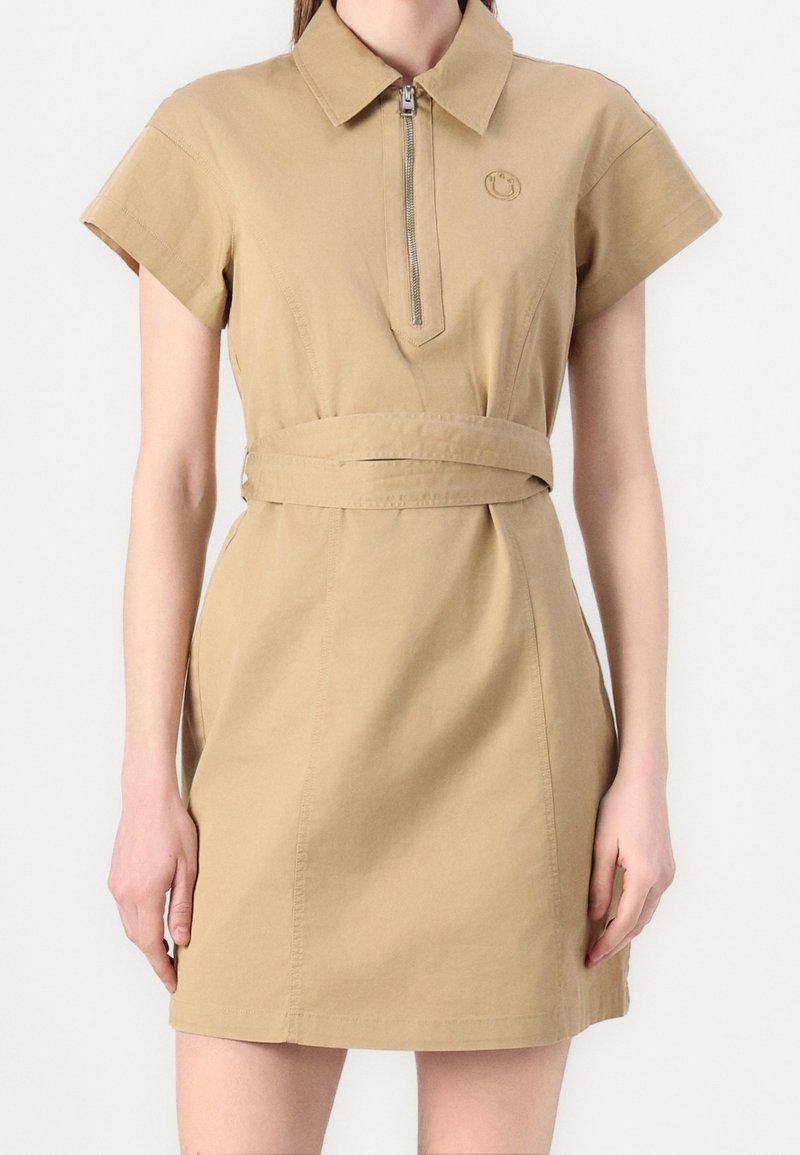 Robe chemise beige à manches courtes avec col, fermeture éclair devant et ceinture à la taille portée par une personne debout devant un fond uni.