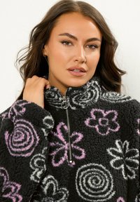 Czarny polarowy pulower z różowymi i białymi wzorami kwiatowymi oraz spiralnymi, z wysokim kołnierzem i zapięciem na suwak z przodu. Miękka, teksturowana tkanina.