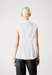 Bluză albă fără mâneci, cu guler croit, având o siluetă ajustată și pliuri subtile în spate, completată cu pantaloni gri închis.