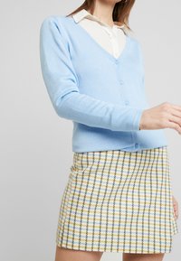 Světle modrý bavlněný cardigan s výstřihem do V a knoflíkovým zapínáním, doplněný o žlutou a modrou kostkovanou mini sukni. Hladká textura.