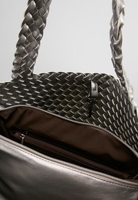 Borsa intrecciata in argento con manici intrecciati, caratterizzata da un interno marrone e una tasca con cerniera. Esterno liscio con texture metallica.