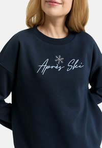 Navy blue sweatshirt with ribbed crew neck, relaxed fit, featuring embroidered "Après Ski" text and a snowflake detail.