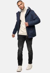 Marineblauwe geïsoleerde jas met een capuchon, gewatteerde voering en voorzakken. Draag het over een lichtbeige coltrui en combineer het met donkere jeans en laarzen.