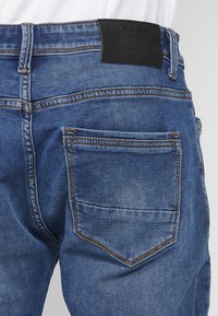 Jeans de denim azul con una superficie texturizada, que presentan costuras naranjas, un bolsillo trasero y una etiqueta de cuero negro en la cintura.