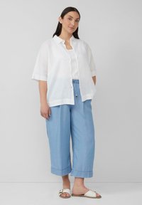 Chemise en lin blanche à manches courtes, associée à un pantalon large bleu et des sandales blanches à glisser avec un accent doré. Look décontracté, coupe ample.