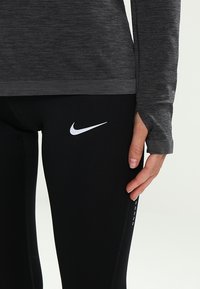 Černé legíny s bílým logem Nike a dlouhým šedým topem s texturou, který má otvory pro palce a žebrovaný lem.