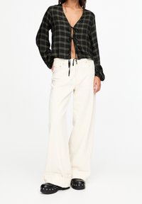 Blusa corta a quadri nera e verde con chiusura a lacci, abbinata a jeans bianchi a gamba larga e scarpe platform nere con borchie silver.