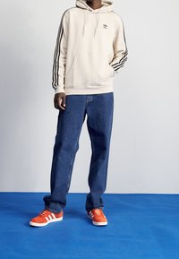 Beige huvtröja med svarta ränder, broderad logotyp, framficka; blå vida jeans; orange sneakers med vita ränder.