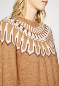 Pull en maille brun clair avec un motif géométrique blanc et brun foncé sur le haut de la poitrine et les épaules, porté par une personne aux cheveux blonds.