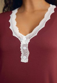 Bordo top s bílou krajkovou lemovkou kolem výstřihu do V a se třemi bílými knoflíky. Měkká tkanina s hladkým povrchem.
