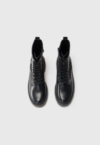 Une paire de bottes de combat en cuir noir à lacets avec des semelles épaisses, vues d'en haut sur un fond clair.