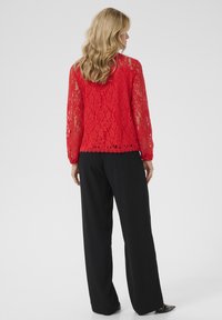 Red lace top with long, sheer sleeves, featuring a scalloped hem. Paired with loose black pants. Background is plain white.