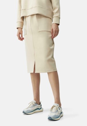 Henkilö pukeutunut beigeen polvipituiseen hameeseen, jossa on etuhalkio ja ylisuuret taskut, yhdistettynä vaaleanharmaisiin ja valkoisiin tennareihin.