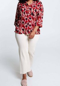 Donna che indossa una blouse rossa a fantasia, pantaloni bianchi e mocassini rosa chiaro su uno sfondo bianco puro.