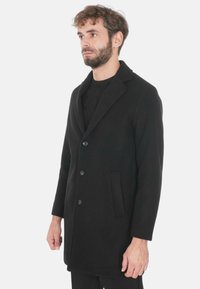 Cappotto di lana nera con rever a lappola, tre bottoni frontali e tasche laterali. Tessuto liscio e design fino al ginocchio.