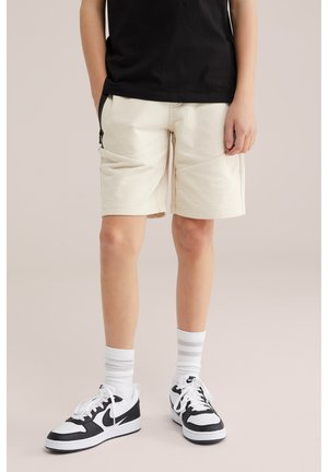 Personne portant un short beige, un t-shirt noir, des baskets blanches Nike et des chaussettes blanches montantes avec des rayures grises, debout devant un fond uni.