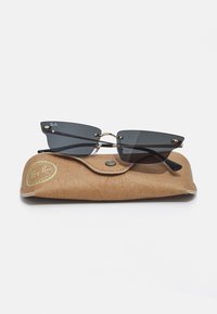Ray-Ban saulesbrilles ar melniem stikliem un stūrainām rāmītēm, novietotas uz brūnas ādas futlāra ar zelta logo un teksturētu virsmu.