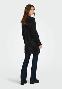 Cardigan long en tricot noir à texture douce, associé à un jean évasé foncé et des chaussures noires épaisses. Le modèle est tourné de dos à la caméra.