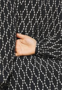 Schwarzer Stoff mit einem wellenförmigen weißen Punktmuster; hat einen Ärmel mit Umschlag und gerafftem Textur. Eine Hand befindet sich unter dem Stoff.