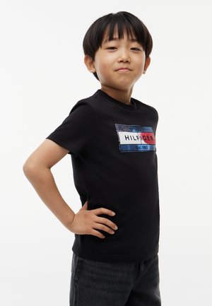 Junge mit kurzen schwarzen Haaren steht selbstbewusst mit den Händen in den Hüften und trägt ein schwarzes T-Shirt mit einem bunten rechteckigen Logo.