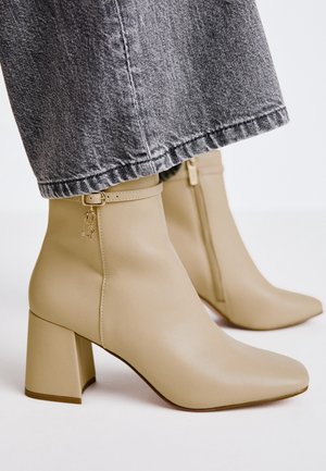 Botas de tobillo de cuero beige con punta cuadrada, tacón de bloque, cremallera lateral y una correa decorativa con detalle de herrajes. Combinadas con jeans gris.