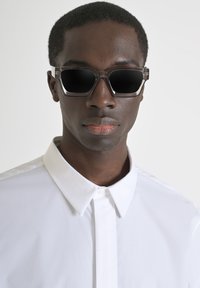 Schwarze Sonnenbrille mit quadratischen Gläsern, transparenten grauen Rahmen und dezenten metallischen Akzenten. Tragen mit einem weißen Hemd mit Kragen.