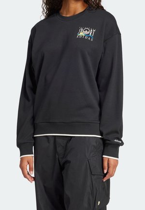 Persoon draagt een zwarte Adidas-trui met kleurrijk "Sport Originals Adidas"-logo op de borst en zwarte cargobroek met zijzak.