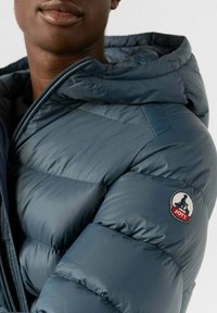 Chaqueta acolchada azul oscuro con capucha, que presenta tela brillante, textura acolchada y una insignia con logo circular en el hombro izquierdo. Detalle de cremallera visible.