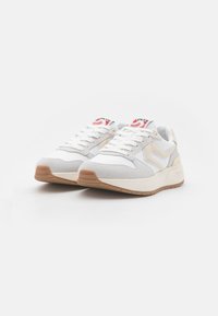 Baskets blanches de sport avec des accents en suède gris, présentant une semelle en caoutchouc texturée, un design à lacets et un logo rouge et bleu sur la languette.
