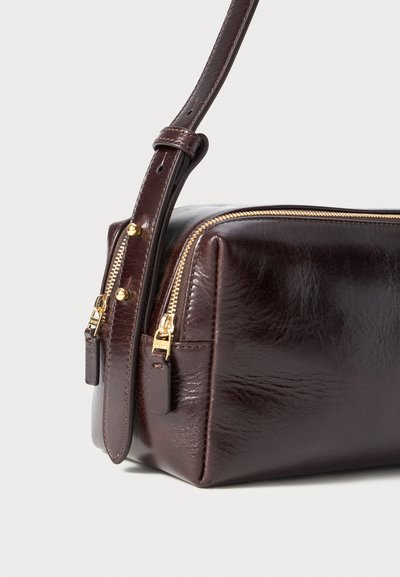 Καφέ δερμάτινη τσάντα με ορθογώνιο σχήμα, με χρυσό φερμουάρ και ρυθμιζόμενο λουρί, που παρουσιάζει λεία υφή και διακριτή λάμψη.