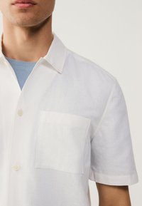 Bijela košulja kratkih rukava od laganog materijala, s jednim džepom na prsima, ovratnikom na kopčanje i teksturiranim završetkom.