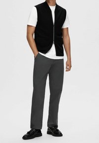 Svart stickad väst med dragkedja, bärs över en vit t-shirt. Kombineras med mörkgrå byxor och svarta texturerade skor.