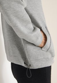 Šedá mikina se boční kapsou, texturovaná tkanina a šňůrka v dolním lemu. Vlastnosti zahrnují volný střih a kontrastní stehy.