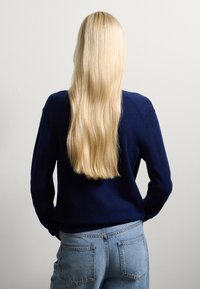 Maglione blu navy realizzato in materiale morbido, abbinato a jeans in denim azzurro. Lunghi capelli biondi lisci scendono lungo la schiena.